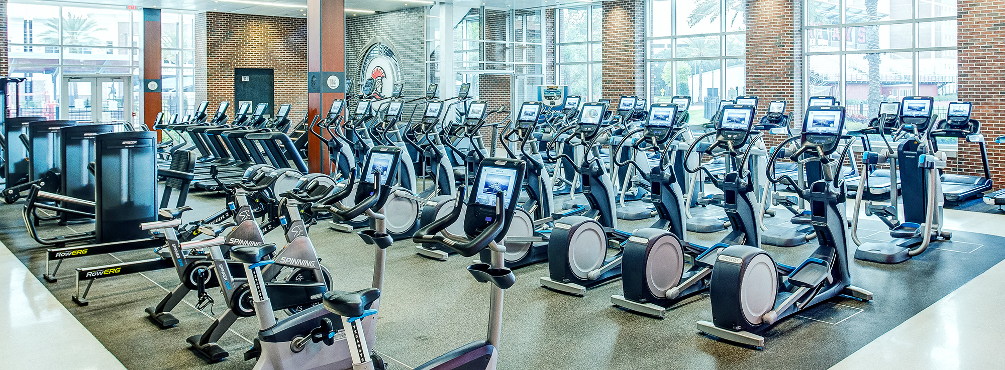 Fitness center with rows of treadmills and ellipticals near large windows and brick walls; bright, modern gym interior